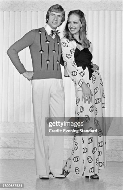 Two fashion models, the man wearing white trousers and a patterned v-neck jumper with a bow tie, and the woman wearing a long skirt and matching...