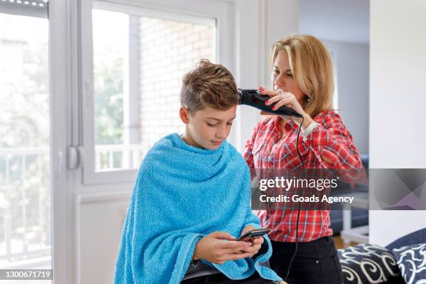 young boy using mobile phone when getting a new haircut from his mother at home - hair clipper stock pictures, royalty-free photos & images