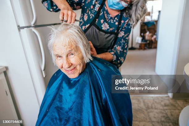 smiling elderly senior caucasian woman getting a haircut at home by a professional caregiver stylist wearing a blue face mask - cutting hair stock pictures, royalty-free photos & images