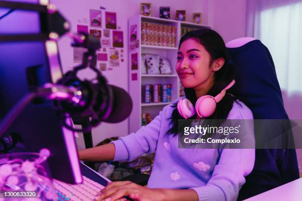 excited and smiling gamer girl in cute headset with mic playing an online video game. young asian woman talking to players and audience on personal computer at home - mood stream stock pictures, royalty-free photos & images