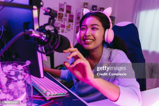 excited and smiling gamer girl in cute headset with mic playing an online video game. young asian woman talking to players and audience on personal computer at home - live streaming stock pictures, royalty-free photos & images