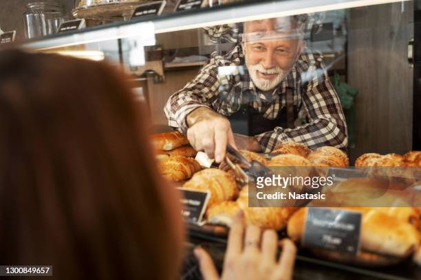 senior bagare man i bageributik välja bakverk med servering tång för en kund - artisan food bildbanksfoton och bilder