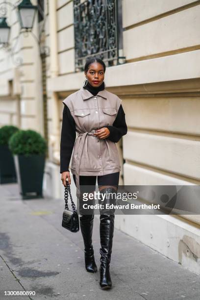 Magalie Kab wears large earrings, a black wool turtleneck pullover from Zara, a sleeveless gray leather jacket/short dress, short leggings, thigh...