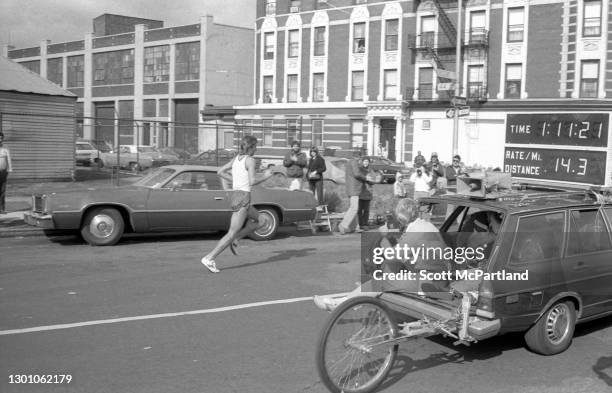 View of an unidentified lead runner along side the official pace car featuring their time on the race, as they pass the 14.3 mile marker, on Crescent...