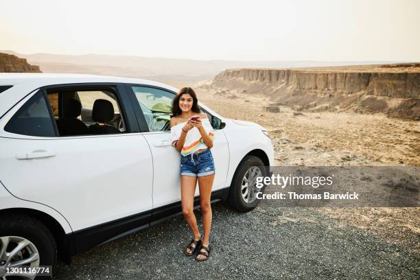 smiling teenage girl holding smart phone and hanging out next to car during family road trip - american girl alone stock pictures, royalty-free photos & images