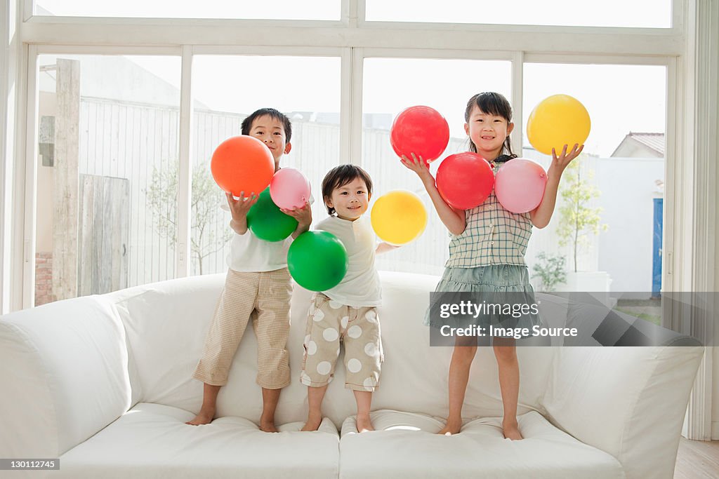 Three children standing on sofa with balloons