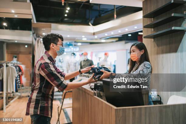 asian chinese beautiful female menswear store business owner cashier with face mask receiving clothes purchased by her customer putting them in bags at cashier counter - small business mask stock pictures, royalty-free photos & images