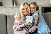 Surprise for mother's day or birthday. Loving young adult daughter giving a present to her beloved middle-aged caucasian mom sitting on the sofa in the living room