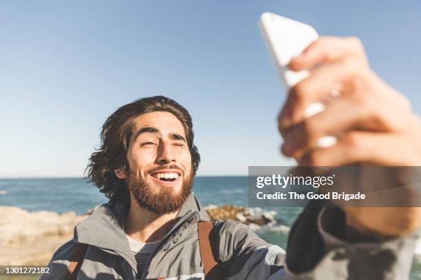 cheerful young man taking selfie through smart phone at beach - selfie male stock pictures, royalty-free photos & images