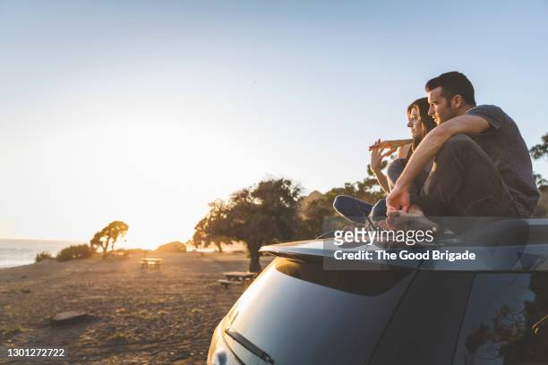 young couple sitting on top of car while looking at sunset - sitting on top of car stock pictures, royalty-free photos & images