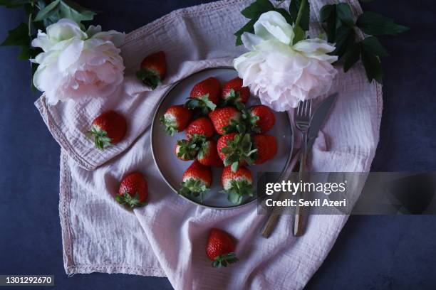 frische erdbeeren auf einem grauen teller auf einem dunkelblauen steinboden, eine rosa stoffserviette mit spitze am rand unter dem teller, verziert mit zwei großen künstlichen rosa puoniden, erdbeeren verstreut, ein besteck neben dem teller - food table edge stock-fotos und bilder