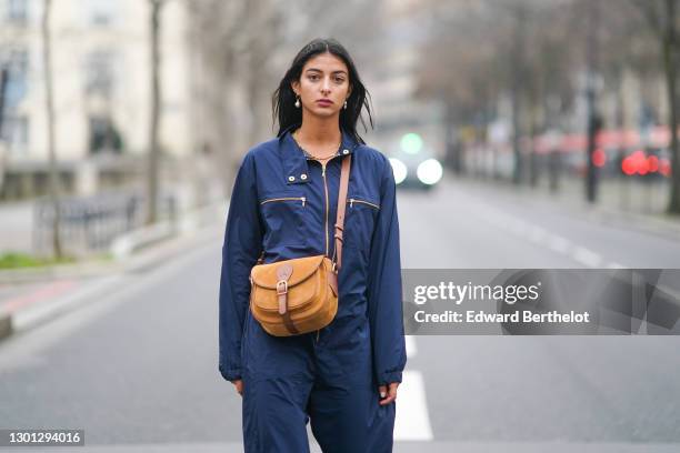 Melanie Darmon wears pearl earrings, a blue jumpsuit, a brown suede bag with leather parts from Gérard Darel, gray wool socks, on February 09, 2021...