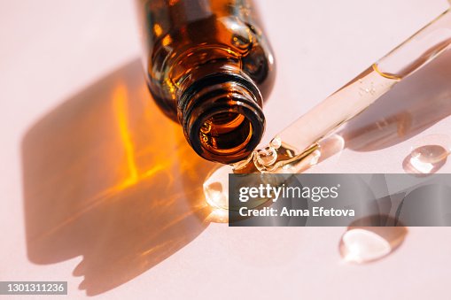 Drops of facial serum or polyglutamic acid pouring out of amber glass bottle nearby pipette on pink background. Trendy products of the year. Health and wellness concept Drops of facial serum or polyglutamic acid pouring out of amber glass bottle nearby pipette on pink background. Trendy products of the year. Health and wellness concept