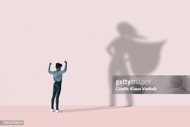 woman flexing muscles in front of superhero shadow - obiettivo foto e immagini stock