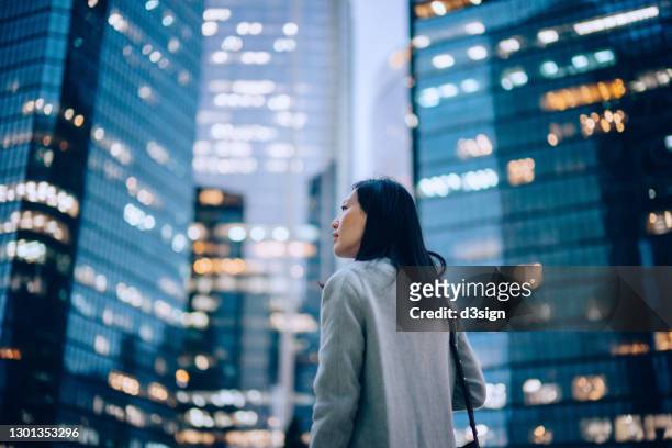 low angle side profile of confident and professional young asian businesswoman looking up while standing against contemporary corporate skyscrapers with illuminated facade in financial district in the evening. female leadership and determined to success - corporate business photos et images de collection