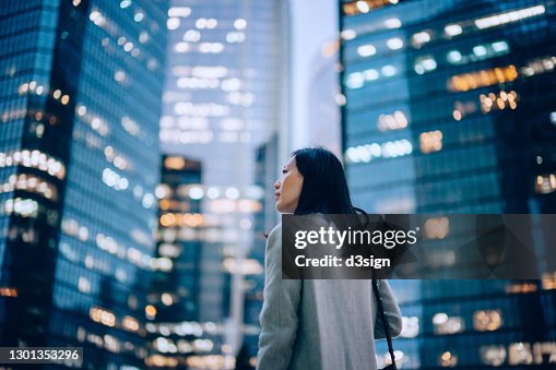 Low angle side profile of confident and professional young Asian businesswoman looking up while standing against contemporary corporate skyscrapers with illuminated facade in financial district in the evening. Female leadership and determined to success Low angle side profile of confident and professional young Asian businesswoman looking up while standing against contemporary corporate skyscrapers with illuminated facade in financial district in the evening. Female leadership and determined to success