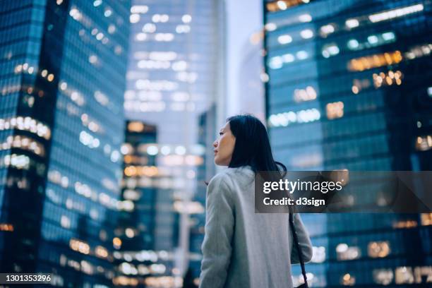 low angle side profile of confident and professional young asian businesswoman looking up while standing against contemporary corporate skyscrapers with illuminated facade in financial district in the evening. female leadership and determined to success - point of view stockfoto's en -beelden