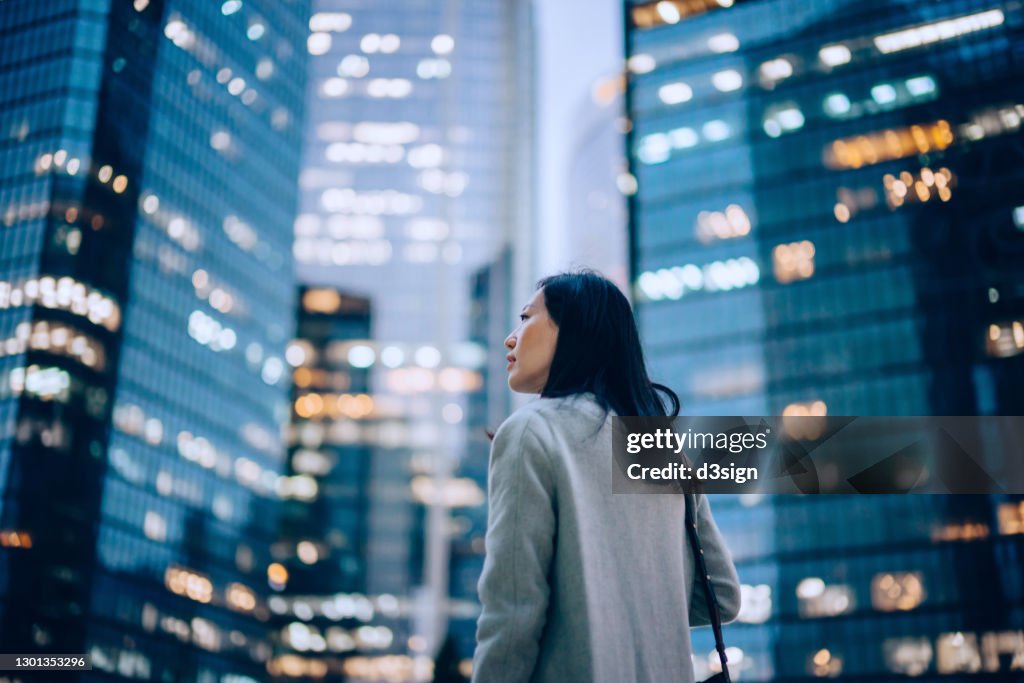 Low angle side profile of confident and professional young Asian businesswoman looking up while standing against contemporary corporate skyscrapers with illuminated facade in financial district in the evening. Female leadership and determined to success