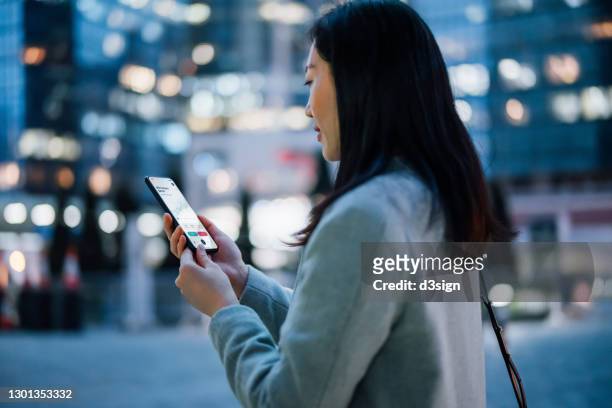young asian businesswoman checking financial stock market analysis with mobile app on smartphone on the go, against illuminated contemporary corporate skyscrapers in financial district in the evening. business on the go - blockchain crypto stock pictures, royalty-free photos & images