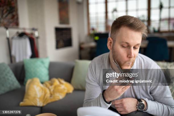 young man sitting on a sofa and trimming his beard while relaxing at home - beard trimming stock pictures, royalty-free photos & images