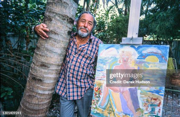 American playwright Tennessee Williams hugging a palm tree trunk while posing for a portrait next to an oil painting on an easel depicting a nude...