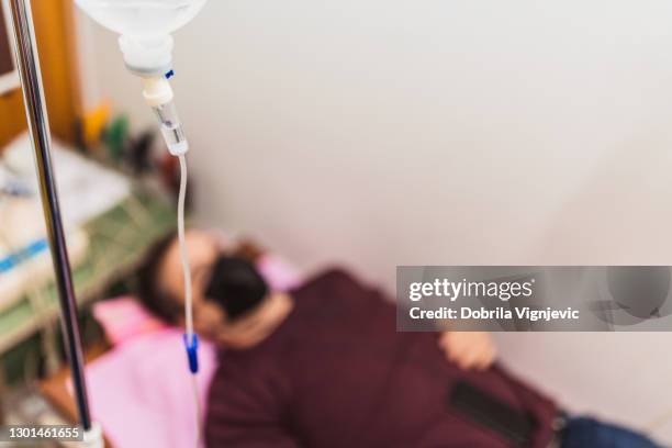 close-up tiro de um gotejamento intravenoso com um paciente inconsciente no fundo - infused - fotografias e filmes do acervo