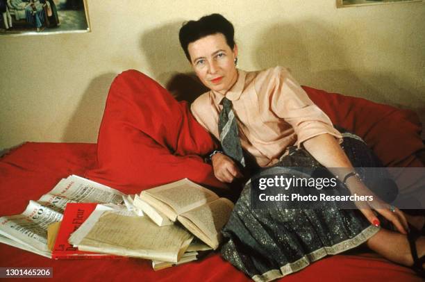 Portrait of French writer and philosopher Simone de Beauvoir as she reclines on a red couch, several newspapers and books in front of her, Paris,...