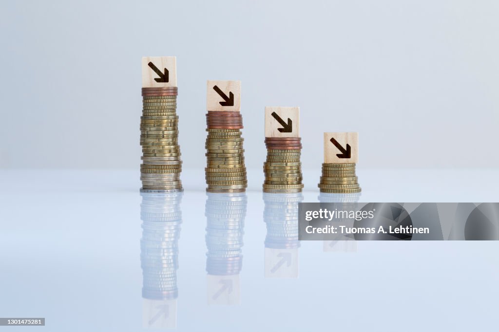 Arrows on wooden blocks on top of descending stacks of coins against gray background.