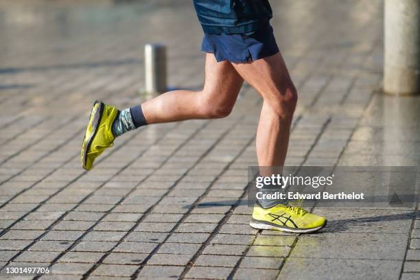 Passerby wears neon yellow Asics running sneakers shoes, socks, blue sport shorts, on February 10, 2021 in Paris, France.
