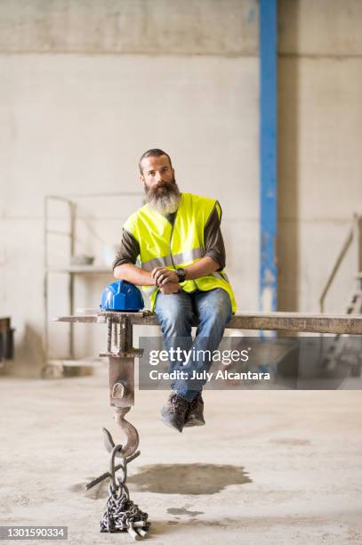bärtiger bauarbeiter ruht auf dem arm eines gabelstaplers, der eine pause von seiner arbeit einlegt - crane hook stock-fotos und bilder
