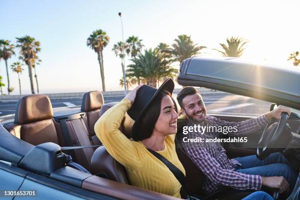 joven sosteniendo su sombrero mientras conduce en un coche convertible con su novio - convertible fotografías e imágenes de stock