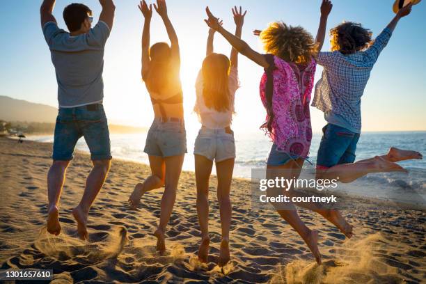 group of friends jumping for joy on the beach at sunset or sunrise. - lens flare young people dancing on beach stock pictures, royalty-free photos & images