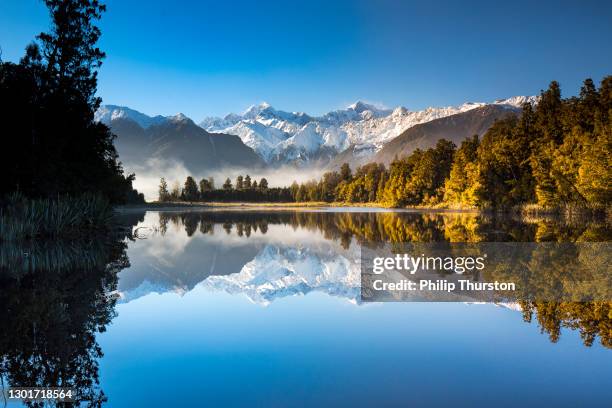 golden snow capped mountain range reflecting perfectly on mirror lake with bright blue sky - lake matheson new zealand stock pictures, royalty-free photos & images
