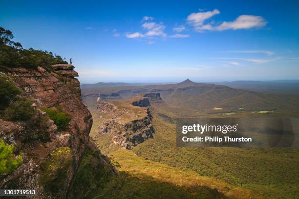 man standing on edge of cliff overlooking vast mountain ranges and valleys on sunny day - blue mountain stock pictures, royalty-free photos & images