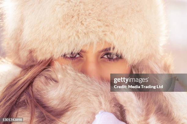 mooie vrouw met bonthoed die camera in het winterpark in de stad tijdens de dag in sneeuwweer met dalende sneeuw, close-up bekijkt - bontmuts stockfoto's en -beelden