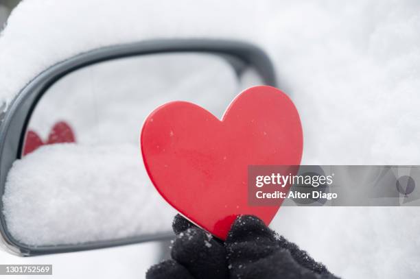 a person holds a red heart shape in front of a rear view mirror. of a car covered with snow - frostbite fingers stock-fotos und bilder