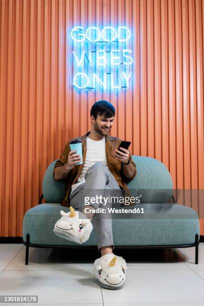 smiling creative businessman with coffee cup in unicorn slippers using smart phone against wall in office - business shoes stockfoto's en -beelden