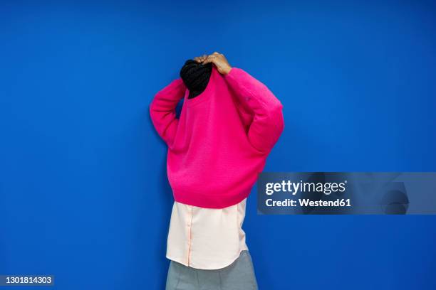 female entrepreneur removing sweatshirt while standing against blue wall at work place - entkleiden stock-fotos und bilder