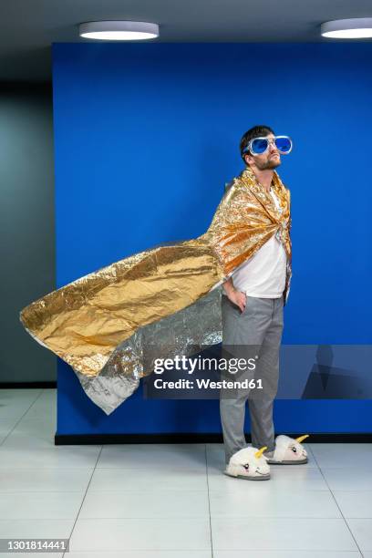 creative businessman wearing cape made of gold color wrapping paper against blue wall in office - funny slipper stock pictures, royalty-free photos & images