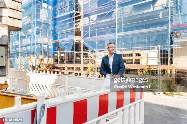 male engineer looking away while standing by barricade at construction site - person in suit construction stock pictures, royalty-free photos & images