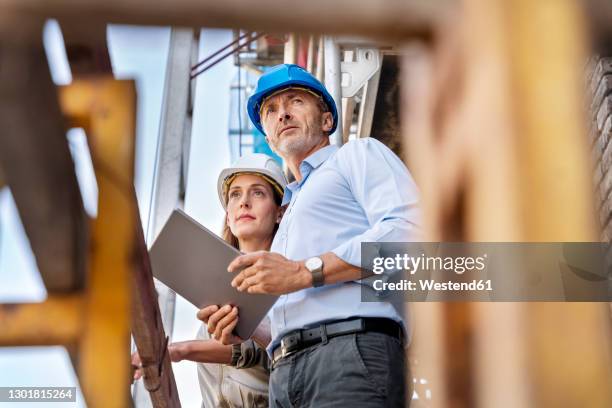 male architect holding digital tablet while examining construction site with coworker - hard hat ipad stock pictures, royalty-free photos & images