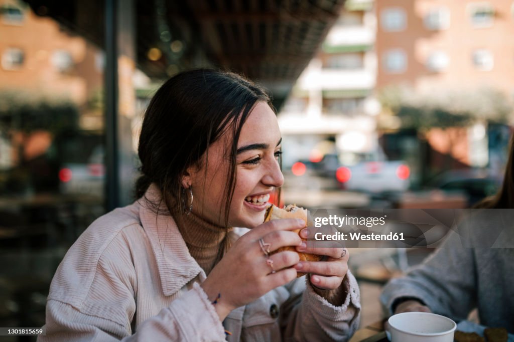 Close-up of cheerful teenage girl eating burger with friend at sidewalk cafe