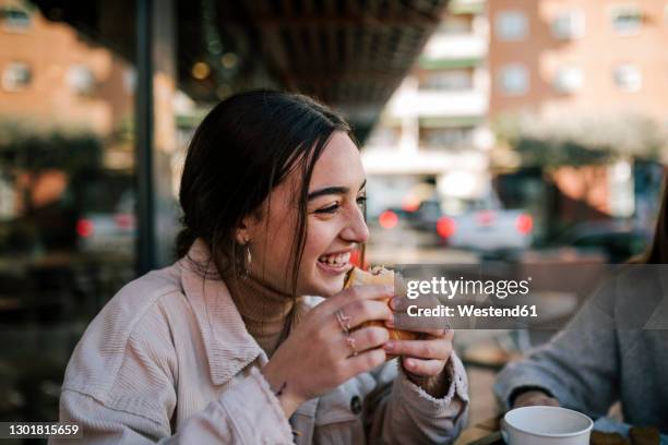 close-up of cheerful teenage girl eating burger with friend at sidewalk cafe - eat stock pictures, royalty-free photos & images