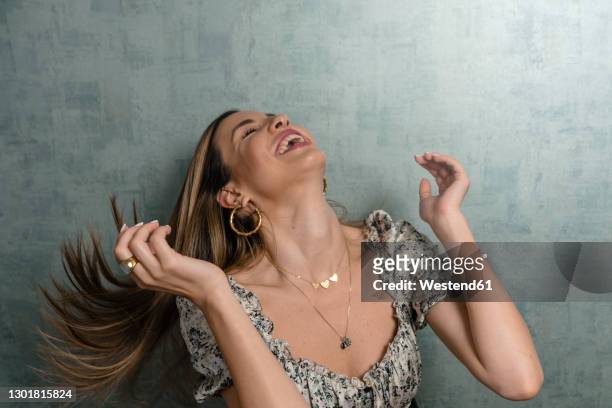 cheerful woman tossing hair while sitting against concrete wall - collana foto e immagini stock