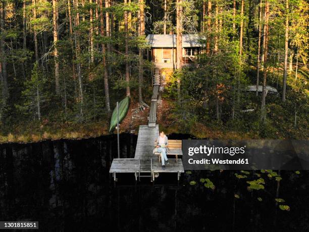 drone shot of woman reading book while sitting on pier over lake in forest - drone images stock-fotos und bilder