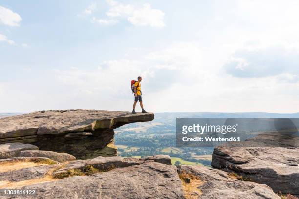 young male hiker looking at view while standing on top of mountain during sunny day - cliff side stock pictures, royalty-free photos & images