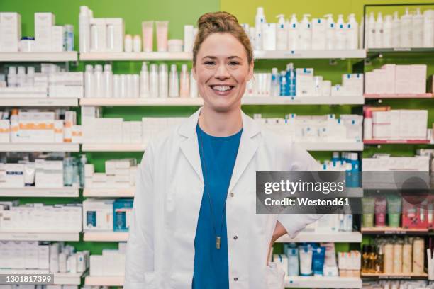 happy pharmacist with hand on hip standing in chemist shop - apothekerin stock-fotos und bilder