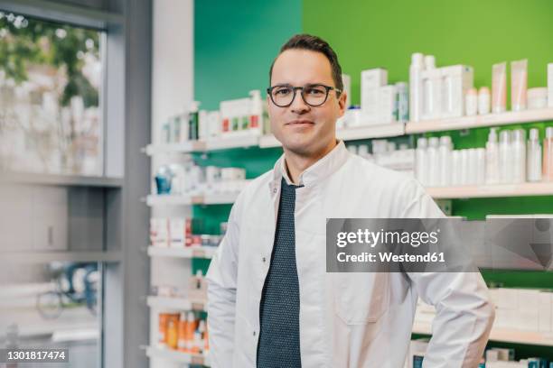 smiling pharmacist wearing lab coat while standing in chemist shop - pharmacist foto e immagini stock