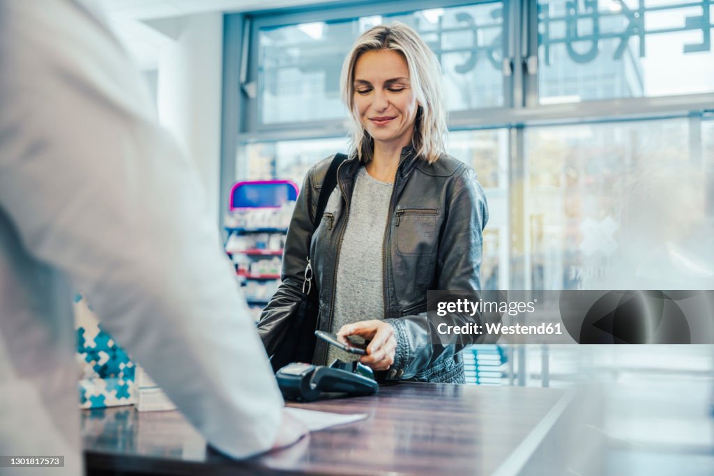 Smiling customer making payment through mobile phone at checkout counter in chemist shop