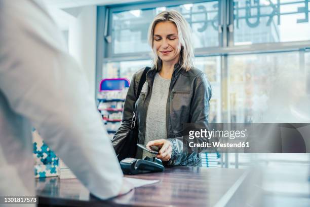 smiling customer making payment through mobile phone at checkout counter in chemist shop - paying stock-fotos und bilder